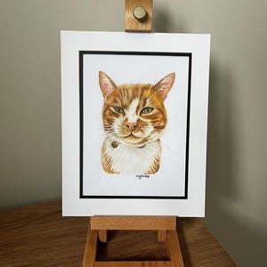 May include: A portrait illustration of an orange and white tabby cat with green eyes, wearing a small collar. The artwork is presented in a white mat with a thin black border, held upright by a light-colored wooden display easel stand.