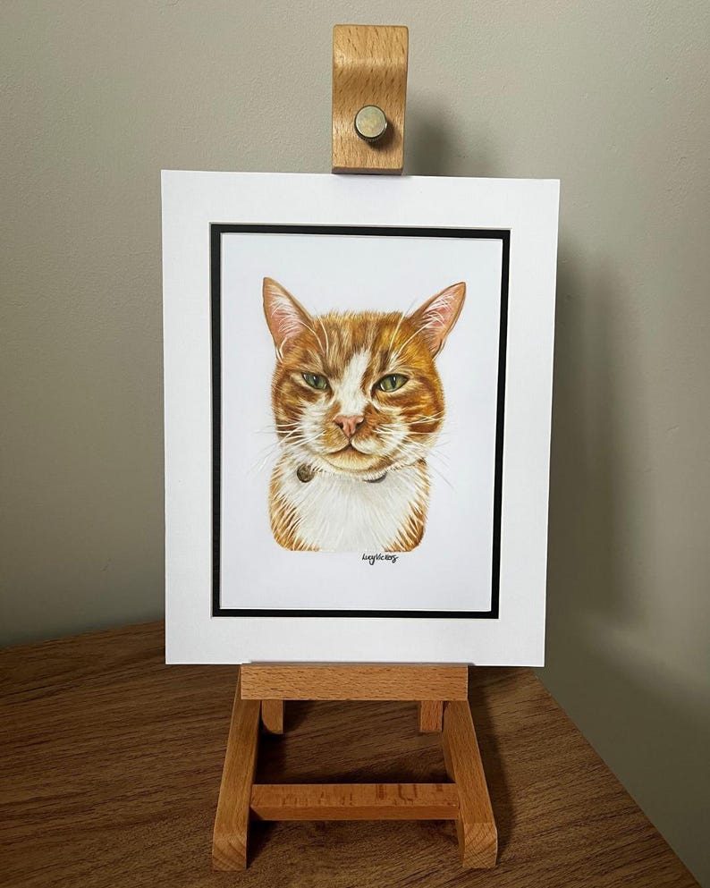 May include: A portrait illustration of an orange and white tabby cat with green eyes, wearing a small collar. The artwork is presented in a white mat with a thin black border, held upright by a light-colored wooden display easel stand.