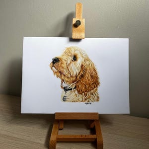 May include: A realistic colored pencil drawing of a golden-colored dog with long, wavy fur, shown in profile facing left. The dog wears a dark collar with a small tag. The artwork is displayed on a small wooden easel against a neutral wall.