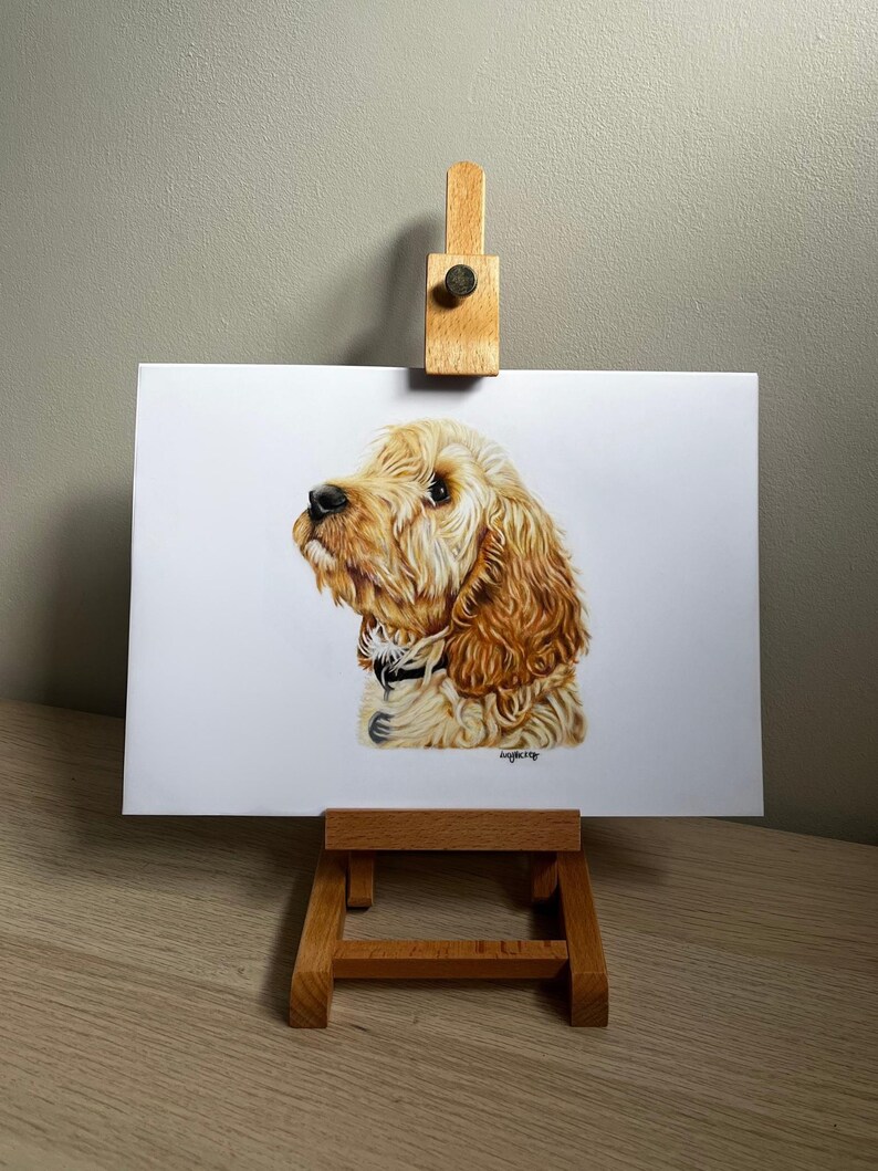 May include: A realistic colored pencil drawing of a golden-colored dog with long, wavy fur, shown in profile facing left. The dog wears a dark collar with a small tag. The artwork is displayed on a small wooden easel against a neutral wall.