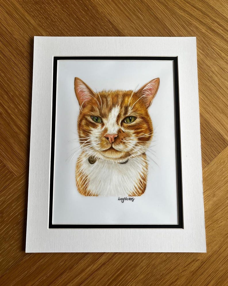 May include: A portrait illustration of an orange and white tabby cat with green eyes, wearing a dark collar. The artwork is set within a black-bordered white matting. The artist's signature, Lucy Vickers, is visible in the bottom right corner.