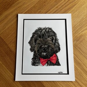 May include: A portrait drawing of a black, shaggy-haired dog wearing a bright red bow tie. The artwork is centered on a white background, framed by a black border and a white matting, with a signature visible in the bottom right corner.