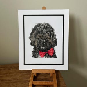 May include: A detailed portrait print of a fluffy black dog with a bright red bow tie, centered within a white mat board featuring a thin black inner border, displayed on a small wooden easel stand for clear viewing of the artwork.