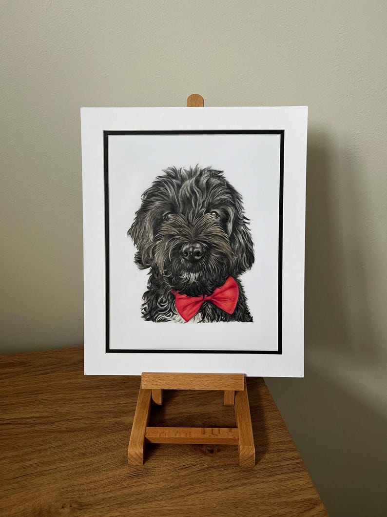 May include: A detailed portrait print of a fluffy black dog with a bright red bow tie, centered within a white mat board featuring a thin black inner border, displayed on a small wooden easel stand for clear viewing of the artwork.