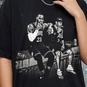 May include: A black short-sleeve t-shirt featuring a monochromatic graphic print of two basketball players sitting on a ledge. One player wears a jersey with the number 23 and the other wears a jersey with the number 3. The shirt has a crew neckline.