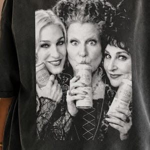 May include: A charcoal gray short-sleeve t-shirt featuring a black and white graphic print of three women holding cans labeled High Noon. The shirt has a crew neckline and a relaxed fit, showcasing a vintage-style wash with a faded appearance.
