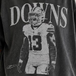 May include: A charcoal gray short-sleeve t-shirt featuring a vintage-style graphic of a football player wearing jersey number 13 with the text DOWNS printed above. The shirt has a distressed, washed-out appearance and a crew neckline.