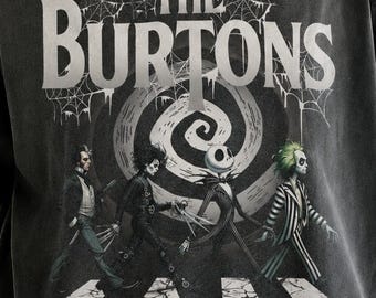 May include: A charcoal gray t-shirt featuring a graphic print of four characters walking across a crosswalk. Above them, the text reads THE BURTONS in white letters, set against a background of spiderwebs and a spiral design. The shirt has a crew neckline.