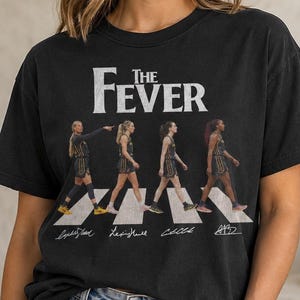 May include: A black short-sleeve t-shirt featuring the text The Fever in white lettering above a graphic of four basketball players walking across a crosswalk. Below the graphic are four white signatures printed on the fabric of the shirt.