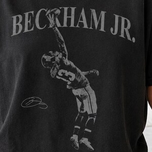 May include: A black short-sleeve t-shirt featuring a faded grey graphic of a football player making a one-handed catch. The text BECKHAM JR. is printed in a large, arched grey font above the player, with a small signature graphic below.