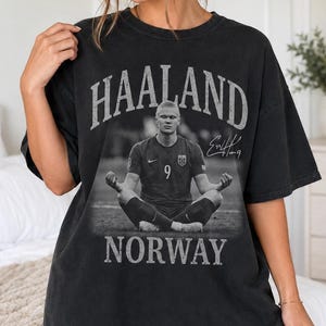 May include: A black short-sleeve t-shirt featuring a graphic print of a soccer player sitting in a meditative pose. The text HAALAND is printed above the image, and NORWAY is printed below. A signature is visible to the right of the player.