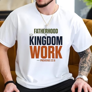 May include: White t-shirt with the text "FATHERHOOD IS KINGDOM WORK" in green, navy, and orange. The text includes a crown graphic and the Bible verse Proverbs 22:6. The shirt is a short-sleeved crew neck.