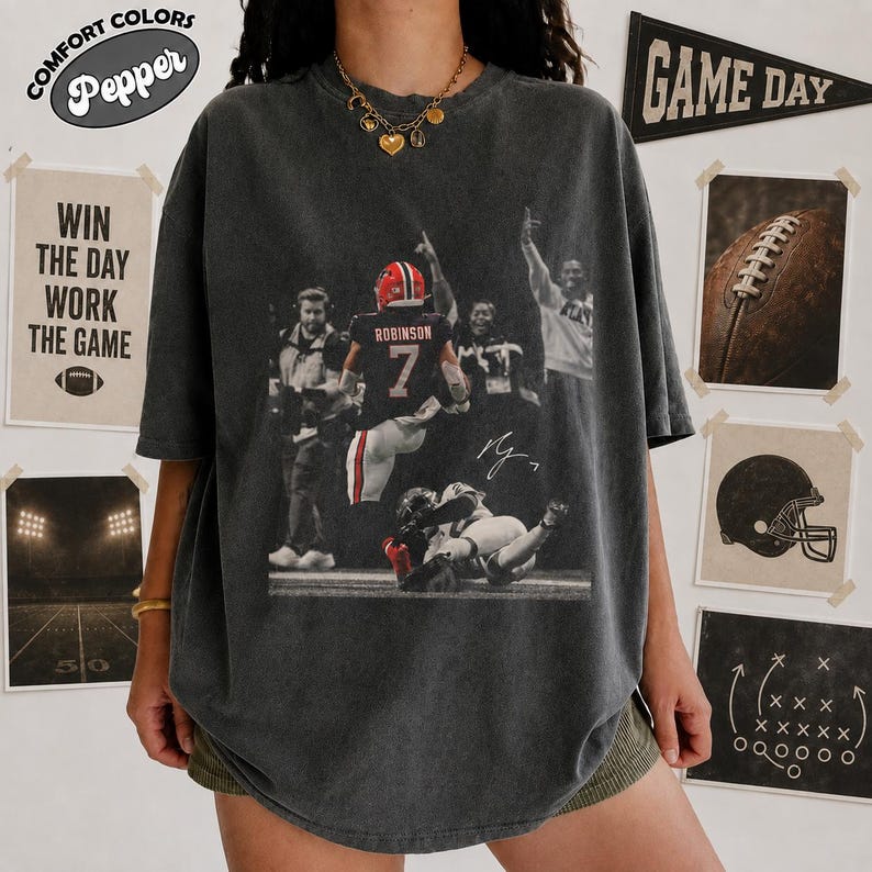 May include: A charcoal grey short-sleeve t-shirt featuring a graphic print of a football player wearing a jersey with the number 7 and the name Robinson. The design includes a black and white action scene of the player on the field.
