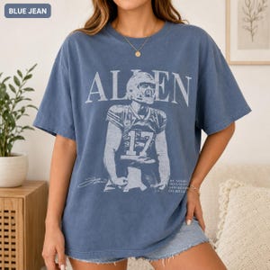 May include: A blue jean colored t-shirt featuring a distressed white graphic of a football player wearing jersey number 17. The text reads ALLEN above the player and BE GOOD GOD BLESS GO BILLS in smaller print on the lower right side.