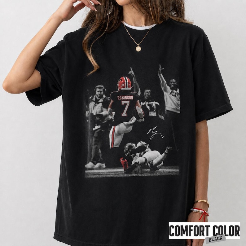 May include: A black short-sleeve t-shirt featuring a graphic print of a football player wearing a jersey with the number 7 and the name Robinson on the back. The graphic includes a signature and depicts a game scene with other figures.