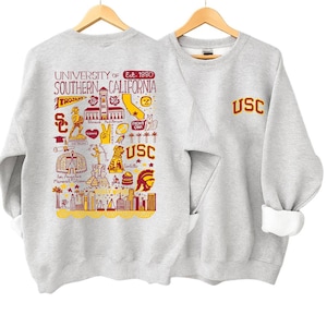 May include: Two light gray sweatshirts hanging on wooden hangers. One sweatshirt features a detailed graphic of Southern California landmarks and the text "UNIVERSITY OF SOUTHERN CALIFORNIA" in maroon and gold. The other sweatshirt has "USC" in maroon.