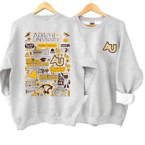 May include: Two light gray sweatshirts are displayed. One features a detailed graphic of Adelphi University, including text and illustrations. The other has the letters "AU" in a gold and brown color. Both are on wooden hangers.