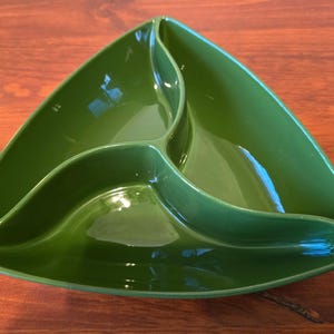 May include: A glossy, green, triangular serving dish with three separate compartments. The dish is made of ceramic or a similar material and sits on a wooden surface. The design is modern and functional, suitable for serving snacks or appetizers.