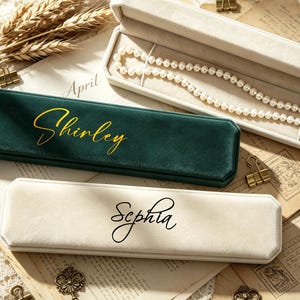 May include: A pearl necklace in an open, off-white jewellery box. Two other velvet jewellery boxes are visible, one green with "Shirley" in gold script and one off-white with "Sephia" in black script. Vintage stationery and gold paper clips are scattered around.