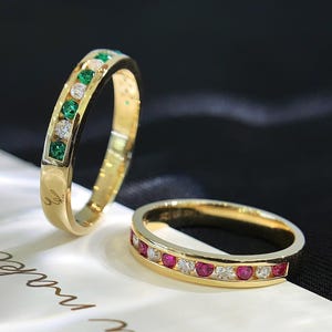 Puede incluir: Dos anillos de oro con piedras preciosas engastadas en canal. Un anillo muestra piedras verdes y transparentes alternadas, mientras que el otro alterna piedras rojas y transparentes. Sobre la superficie blanca aparece el texto You make me.