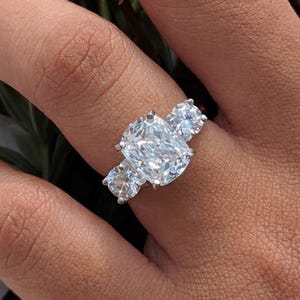 May include: A three-stone ring featuring a large, cushion-cut center diamond flanked by two smaller, round-cut side diamonds. The stones are set in a polished silver-toned metal band, creating a classic and elegant look with clear, faceted surfaces.