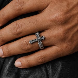 Peut inclure: Une bague en métal argenté présentant un motif de croix proéminent sur le devant. La croix possède une finition texturée et sombre dans ses zones en creux, avec un bord poli. L'anneau de la bague affiche des motifs de volutes gravés.