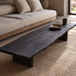 May include: A low-profile rectangular coffee table crafted from dark, charred wood with a visible grain and natural live edge. The table features a flat surface supported by two sturdy block legs, holding a small dark bowl with dried plant stems.