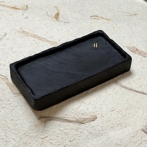 May include: A rectangular black wooden tray with a recessed centre and a rough, textured outer edge. The surface of the tray displays a visible wood grain pattern and features two small, vertical gold-coloured marks near the top right corner.