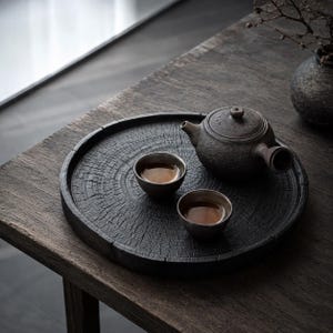 May include: A dark brown, textured ceramic teapot and two matching small tea cups arranged on a circular wooden tray. The tray features a prominent, detailed wood grain pattern resembling a cross-section of a tree trunk with deep, radial cracks.