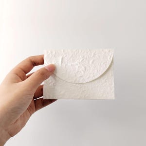 Könnte beinhalten: Kleine, cremefarbene Papierumschlag mit strukturierter Oberfläche. Der Umschlag hat einen gebogenen Klappenverschluss. Der Artikel wird in einer Hand vor weißem Hintergrund gehalten. Die Hülle ist wahrscheinlich für kleine Karten oder Notizen.