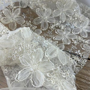 May include: Close-up of white floral lace fabric. The lace features large, three-dimensional flower appliques with delicate petals and silver accents. The background shows more of the same lace, creating a layered effect. This fabric is suitable for bridal wear and other special occasion garments.