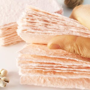 Può includere: Primo piano di pile di carta fatta a mano color pesca con bordi irregolari. La carta ha un aspetto morbido e strutturato. Un dito tocca delicatamente la parte superiore di una pila, evidenziando i delicati strati. La carta viene probabilmente utilizzata per l'arte o l'artigianato.