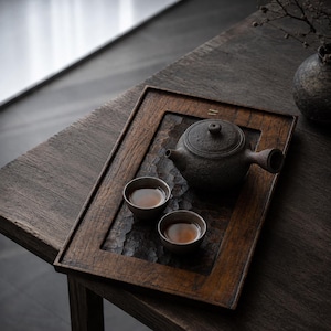 May include: A dark brown rectangular wooden tea tray with a textured surface, featuring a dark gray ceramic teapot with a side handle and two matching small ceramic tea cups filled with amber liquid, arranged neatly on the tray surface.