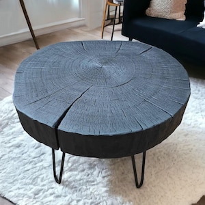Peut inclure: Une table basse ronde dotée d'un plateau épais en tranche de bois gris anthracite, présentant des cernes de croissance naturels visibles et des fissures radiales. Le plateau repose sur quatre pieds fins en épingle en métal noir.