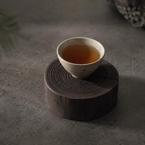 May include: A small, off-white ceramic tea cup filled with amber-colored liquid, resting on a dark, circular wooden log slice that displays visible tree ring patterns and a deep crack across its textured, cylindrical surface.