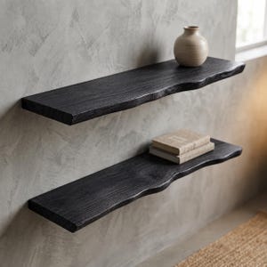 Op de afbeelding: Twee zwarte zwevende houten planken met een natuurlijk levende randontwerp gemonteerd aan een muur. De bovenste plank bevat een kleine, ronde, lichtgekleurde keramische vaas. De onderste plank bevat een stapel van twee boeken met harde kaft.