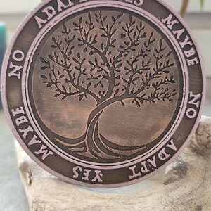 May include: A round, bronze-colored coin with a tree of life design in the center. The coin has a pinkish-brown border with the words "ADAPT YES MAYBE NO" around the edge. The coin is resting on a piece of driftwood.
