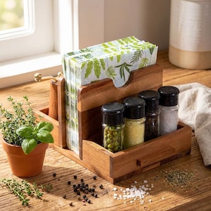 May include: A wooden kitchen organizer with a section for napkins and a compartment for spice jars. The napkins have a green leaf pattern. The spice jars contain green peppercorns, yellow spices, black peppercorns, and salt. A small potted herb plant is also present.