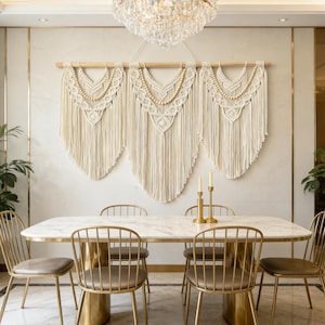May include: A dining room scene featuring a marble-topped table with gold-colored metal chairs. Above the table, three cream-colored macrame wall hangings with wooden beads and fringe are displayed. A crystal chandelier hangs above.