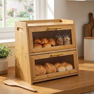 May include: A bamboo bread box with two glass-fronted compartments. The top section holds croissants and cookies, while the bottom contains rolls. Gold-colored handles and a wooden spatula are visible on a wooden countertop.