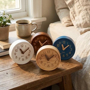 May include: Four small, round analog clocks in white, brown, light wood, and blue. Each clock has a light-colored face with dark brown hands and numbers. The clocks are on a wooden table, with a coffee mug and plant visible in the background.
