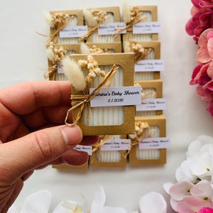 May include: Small, rectangular favor boxes with a window, each containing white items. The boxes are decorated with dried flowers and tied with twine. Each box has a label that reads "Katrina's Baby Shower 2.1.2026".