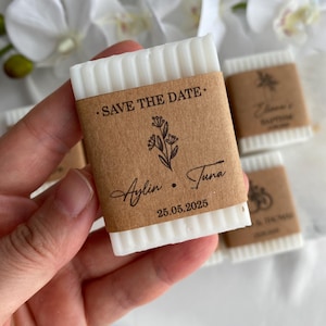 May include: A white, rectangular soap bar with a textured top, wrapped in brown paper. The paper is printed with the words "SAVE THE DATE," a floral design, and the names "Aylin" and "Tuna" along with the date "25.05.2025."