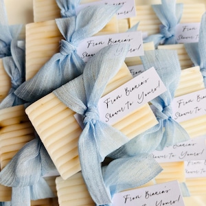 May include: Close-up of several rectangular soaps, each tied with a light blue ribbon and a tag. The tags display the text "From Bianca's Shower To Yours!". The soaps are a light beige colour and have a textured surface.