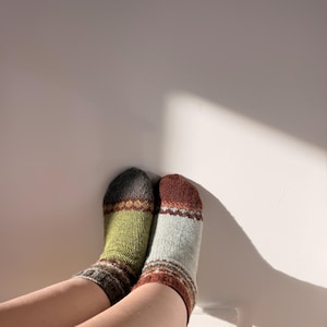 May include: A pair of knitted socks featuring a multi-coloured pattern. The left sock has a dark grey toe, a lime green mid-section, and a dark grey cuff with orange and white accents. The right sock has a rust-coloured toe, a light grey mid-section, and a rust-coloured cuff with white and grey geometric details.