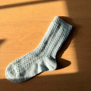 May include: A single light blue knitted sock with a textured cable knit pattern. The sock is lying on a wooden surface, with sunlight casting a shadow across the top. The sock has a ribbed cuff.