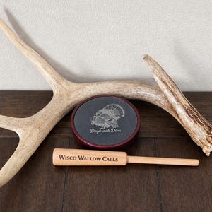 May include: A hunting call set with a wooden striker and a slate call with a turkey graphic. The set includes a large antler and is displayed on a dark wood surface. The text "Daybreak Diva" and "Wisco Wallow Calls" are visible.