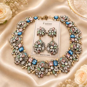 Peut inclure: Un ensemble collier et boucles d'oreilles orné de pierres précieuses colorées. Les bijoux présentent des pierres bleues, roses, vertes et transparentes serties dans un métal doré. Les boucles d'oreilles et le collier sont présentés sur une carte blanche avec "Fashion Jewelry".