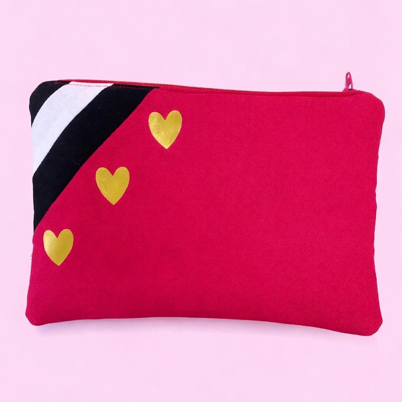May include: A red zippered pouch with a black, white, and black striped corner design. Three gold heart shapes are arranged on the red surface. The pouch is rectangular and has a zipper closure.