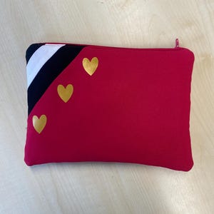 May include: A rectangular red zippered pouch with three gold heart accents. The upper left corner features a black and white striped design. The zipper is red. This pouch is suitable for carrying small items.
